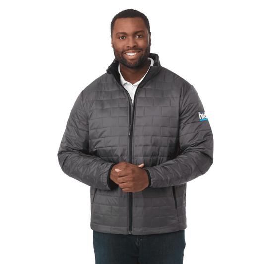 A man wearing a grey puffer jacket with a zip front and a logo on the sleeve.