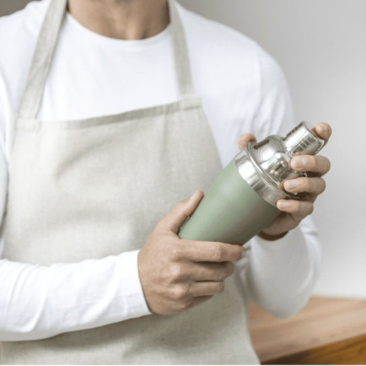 A stainless steel cocktail shaker in matte green, held by a person wearing an apron, with a silver top.