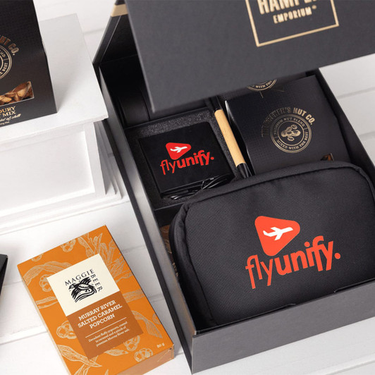 A black gift box contains a branded tech kit, featuring a black pouch, notebook, pen, and snacks.
