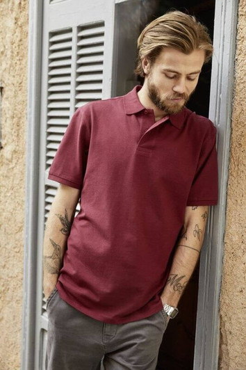 A man with tattoos is wearing a burgundy basic polo shirt while leaning against a doorframe. The shirt has a logo.