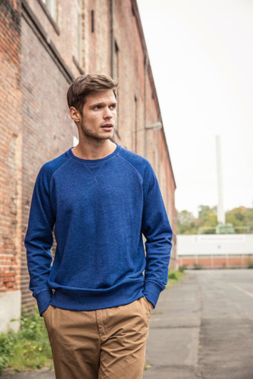 A blue men's jumper is worn by a model standing outdoors near a brick wall, with hands in pockets.