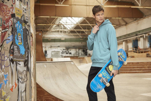 A male model in a blue hoodie holds a skateboard in a skate park setting, with graffiti on the walls.