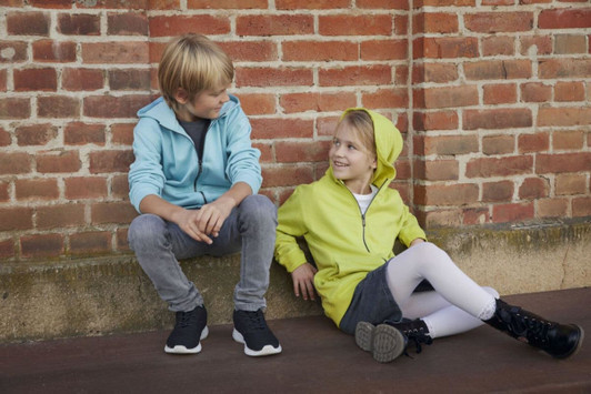 A blue zip hoodie and a lime green zip hoodie worn by a boy and a girl seated against a brick wall.