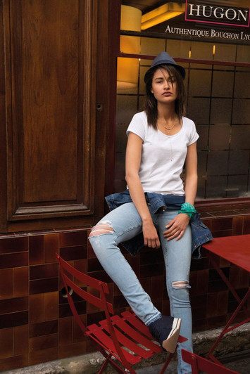 A woman in a white t-shirt and denim jacket sits casually on a red chair, wearing ripped jeans and a grey hat.