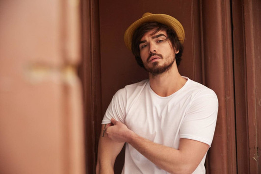 A man wearing a plain white T-shirt and a straw hat, posing against a brown background.