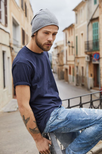 A man wearing a navy blue T-shirt and a grey beanie sits casually on a railing in a street setting.