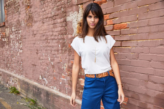 A light-coloured casual T-shirt, styled with blue trousers, worn by a model against a textured brick wall.