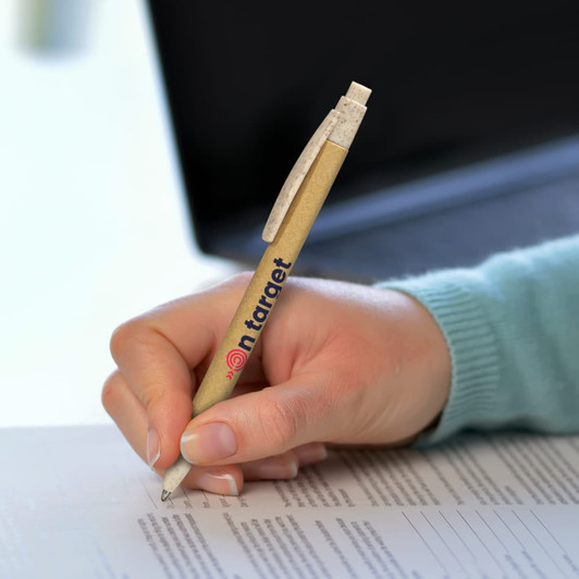 A kraft pen with a natural finish and a clip is held in a hand, writing on a paper document. The pen features a logo.