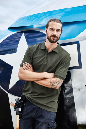 A man in a dark green polo shirt stands with arms crossed beside an aircraft, featuring a logo on the shirt.