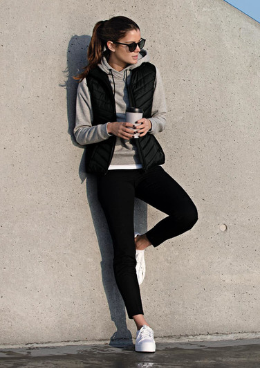 A woman in a black vest over a grey hoodie, holding a drink, leaning against a concrete wall, wearing black leggings and white shoes.