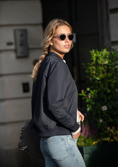 A woman wearing a black puffer jacket and sunglasses, walking past green foliage.