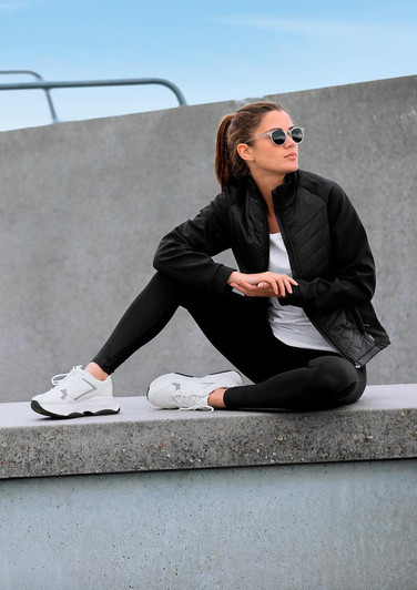 A woman wears a black soft shell jacket with a white top and black leggings, sitting casually against a concrete surface.