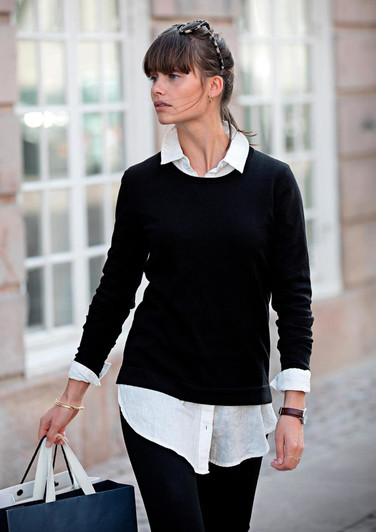 A woman wearing a black knitted jumper over a white shirt, carrying a shopping bag in an urban setting.