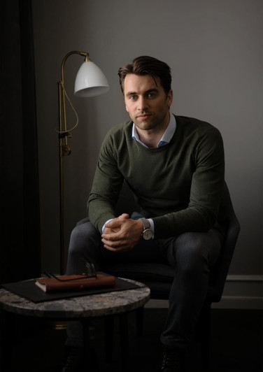 A man sits casually in a chair wearing a green Brighton O-Neck knitwear sweater, with a stylish lamp and table nearby.
