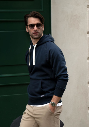 A navy hoodie with a drawstring and front pocket, worn by a man with sunglasses, standing against a green door.