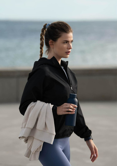 A woman in a black rain jacket carries a drink bottle and a light-coloured sweater, set against a coastal background.