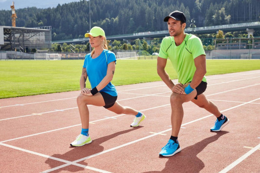 A man in a green sports t-shirt and a woman in a blue sports t-shirt perform lunges on a running track. Both wear caps.