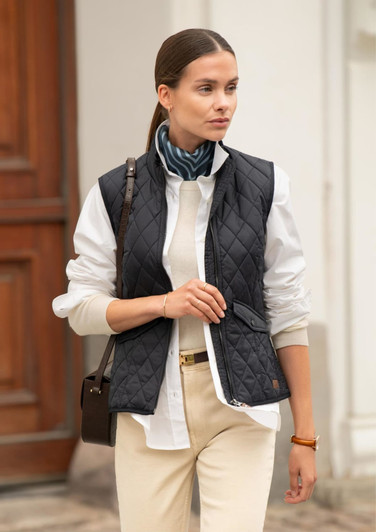 A woman models a navy quilted vest over a white shirt, complemented by a scarf and cream-coloured trousers.