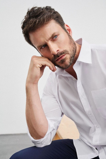 A model in a white long-sleeve button-up shirt, posing thoughtfully with one hand on his chin.