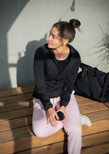 A black soft shell jacket worn by a woman sitting on a wooden bench, paired with light pink pants and a black bag.