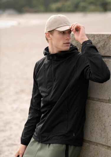 A black soft shell jacket worn by a man, complemented by a beige cap, against a beach backdrop.