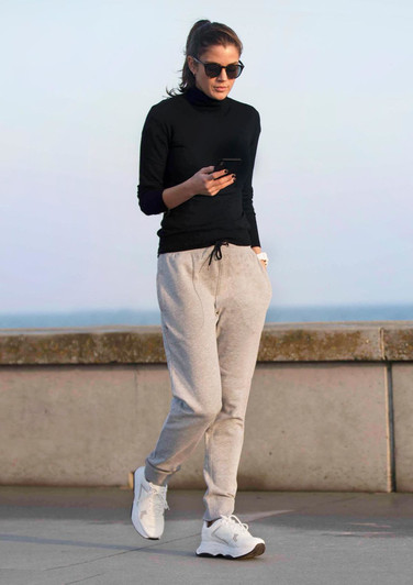 A woman in a black long-sleeve top and grey track pants walks while checking her phone, wearing sunglasses.