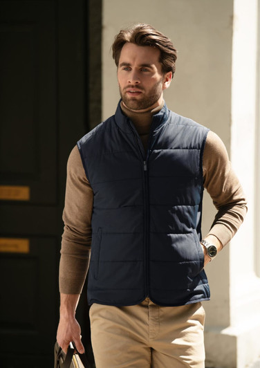 A man wearing a navy blue puffer vest over a tan sweater, walking outdoors. The vest has a zip closure and a logo.
