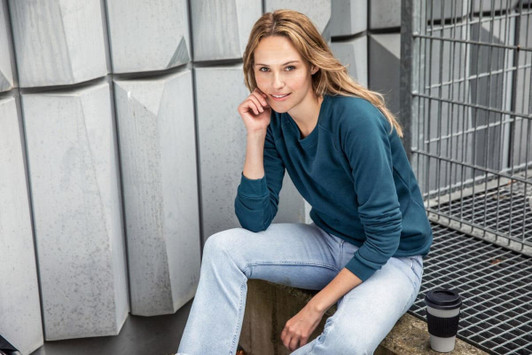 A woman wearing a teal sweater sits casually, with light blue jeans and a coffee cup beside her. The background is textured.