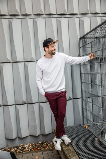 A man is wearing a white jumper and burgundy pants, standing next to a wire fence. He has a black cap on.