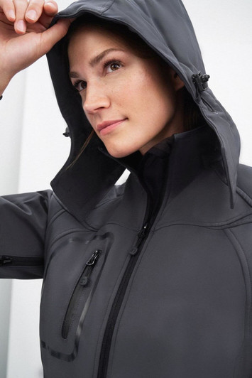 A grey ladies' softshell jacket featuring a hood, with a close-up of a woman wearing it and a logo present.