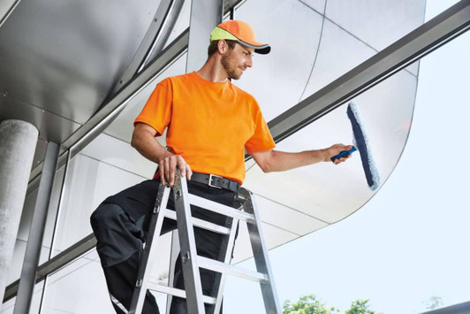 A man in an orange work T-shirt and cap is cleaning windows while standing on a ladder. The setting features modern architecture.
