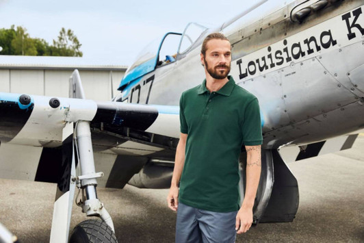 A man stands beside a vintage aircraft, wearing a dark green work polo shirt and grey trousers. The aircraft features a logo.