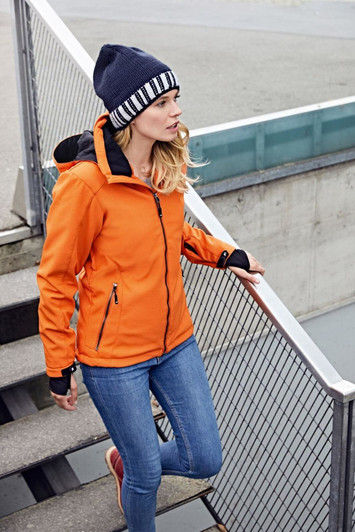 An orange soft shell jacket worn by a woman, paired with a blue beanie and denim jeans, standing on stairs.