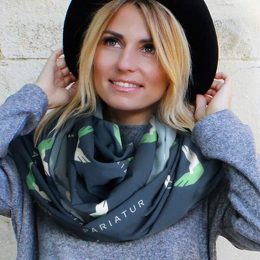 A woman models a grey infinity scarf featuring green hand designs, complemented by a black hat and casual attire.