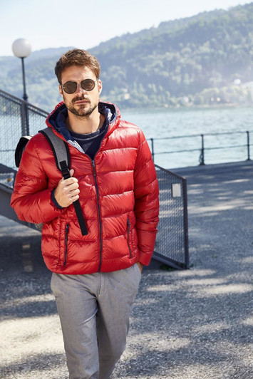 A red men's down puffer jacket with a hood, worn over a navy top, paired with light trousers and sunglasses.