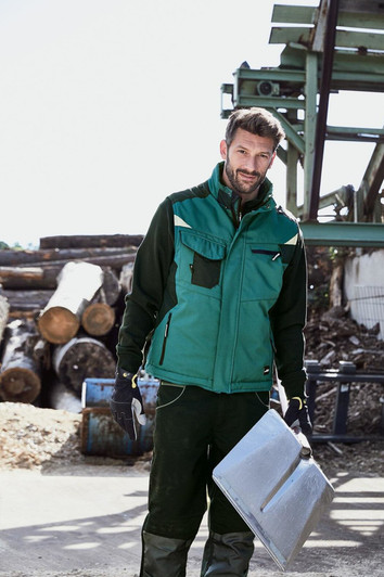 A man stands outdoors wearing a green softshell vest over a black outfit, holding a metal sheet. Logs and machinery are in the background.