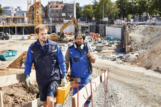 Two men wearing Craftsmen Softshell Vests in blue walk on a construction site with tools in hand.
