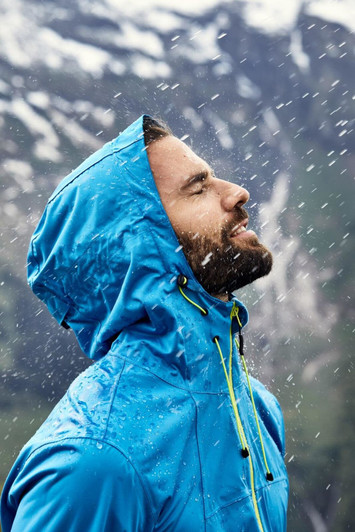 A blue men's outdoor jacket with a hood, worn by a man in the rain against a mountainous backdrop.