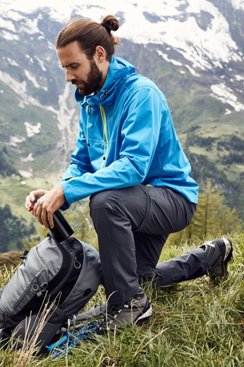A blue men's outdoor jacket is worn by a person kneeling on grass next to a backpack in a mountainous landscape.