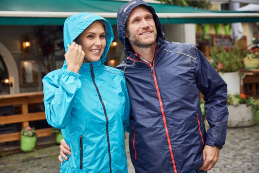 A light blue ladies rain jacket and a navy men's rain jacket, both featuring hoods, in an outdoor setting.