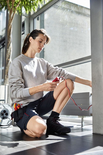 A woman kneels on a tiled floor wearing a grey workwear sweatshirt and black shorts, handling wires with an electrical device nearby.