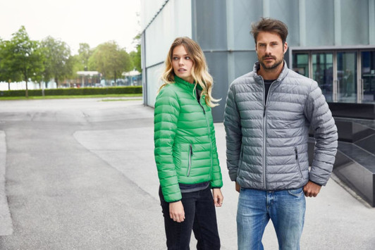 A green puffer jacket worn by a woman and a grey puffer jacket worn by a man stand outside a modern building.