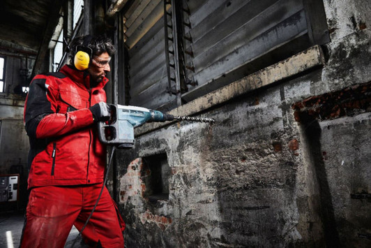 A man in a red workwear softshell jacket is using a power drill in an industrial setting. He is wearing ear protection.