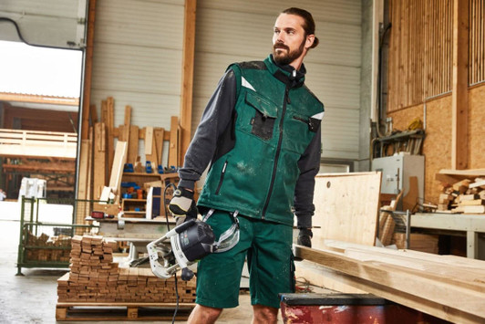 A man wearing a green softshell work vest stands in a workshop, holding a power tool, surrounded by wooden materials.