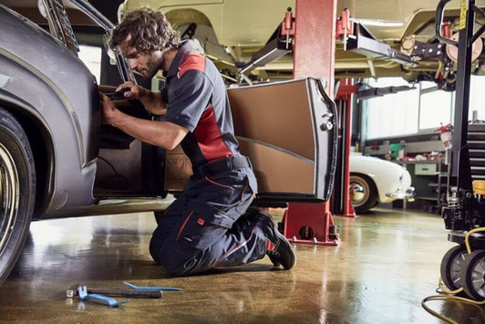 Workwear pants in black with red accents, worn by a person kneeling beside a car in a workshop setting.