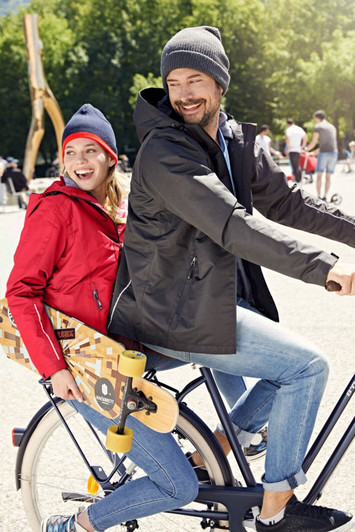 A red and black 3-in-1 jacket worn by a woman and a man on a bicycle, surrounded by greenery.