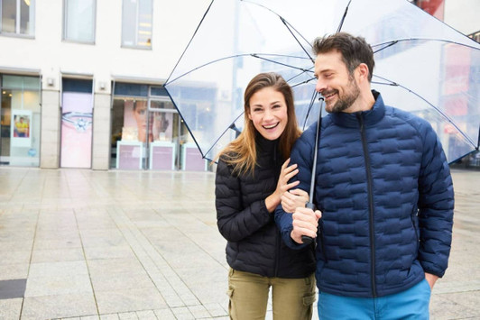 A modern padded jacket in navy, worn by a smiling man and woman under a clear umbrella in a city setting.