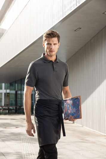 A man in a grey polo shirt and apron holds a menu while walking outside a modern building.
