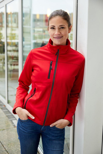 A red ladies softshell jacket with a high collar and zippered pockets, featuring a branding logo.