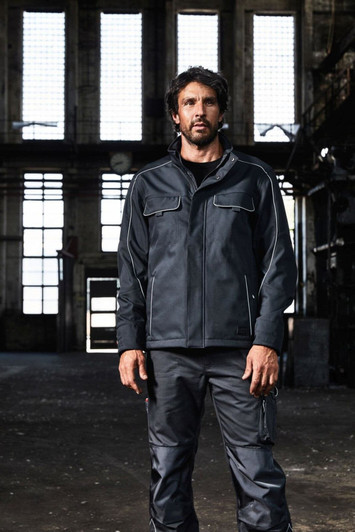 A man wearing dark grey workwear pants and a matching jacket stands in an industrial setting. The image features a logo.